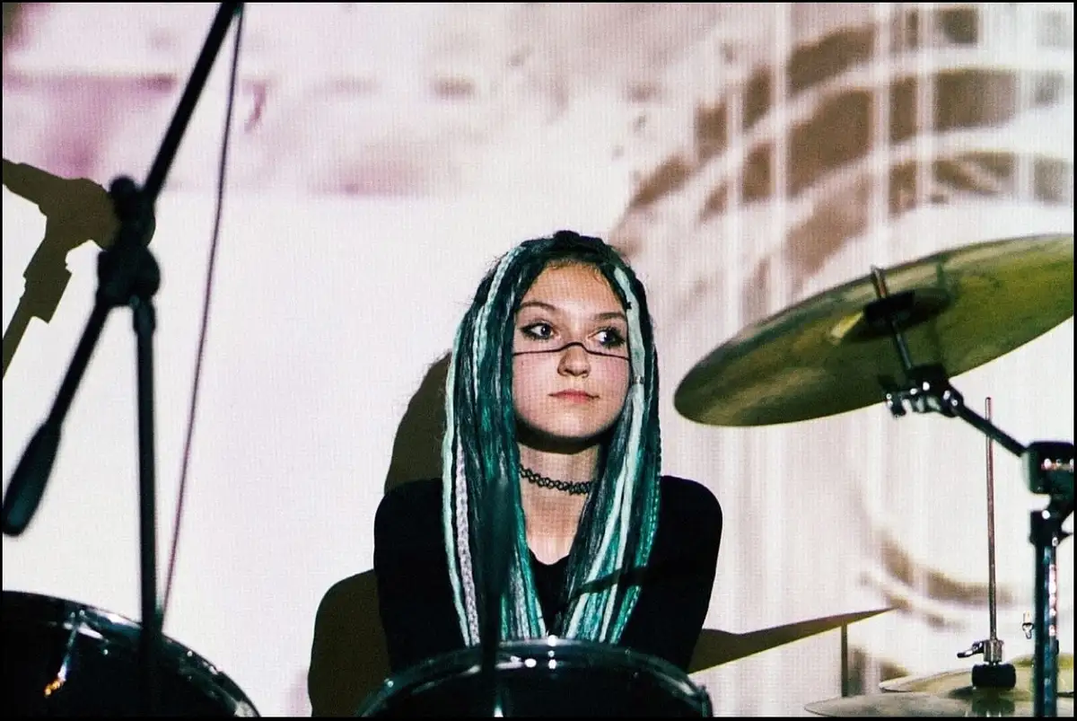 A young woman with teal and black braided hair and glasses sits behind a drum kit, looking intensely at the camera. She…