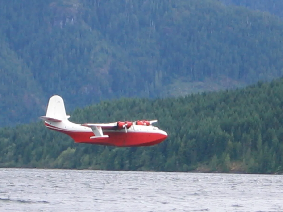 A red and white seaplane with floats is flying low over a body of water, with a dense forested mountain range in the…