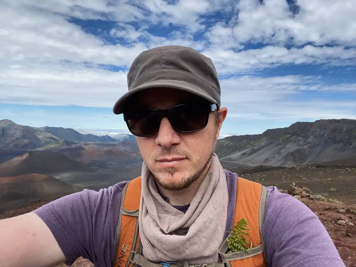 A man wearing a cap, sunglasses, a purple shirt, and an orange backpack stands on a rugged mountain ridge with a vast…