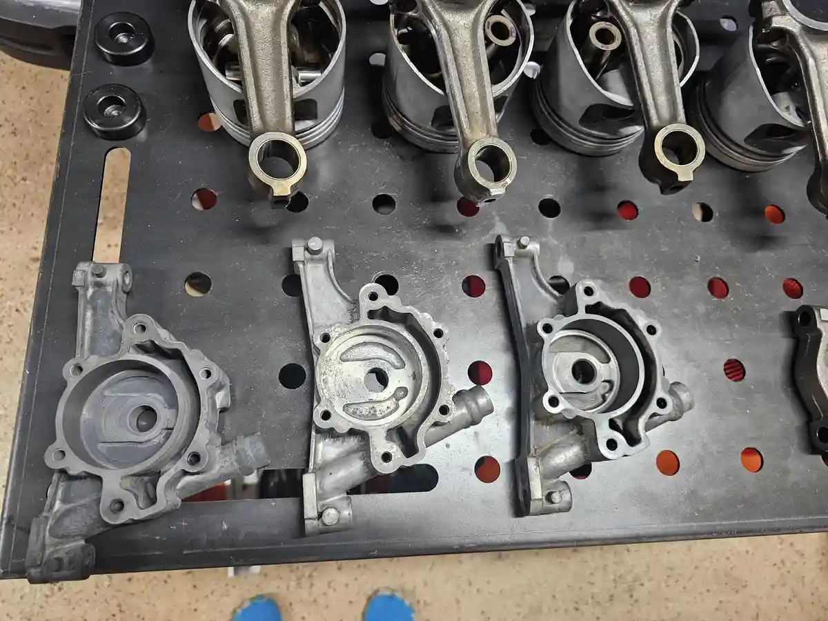 A top-down view of a black perforated metal workbench holding several silver engine components, including pistons and…