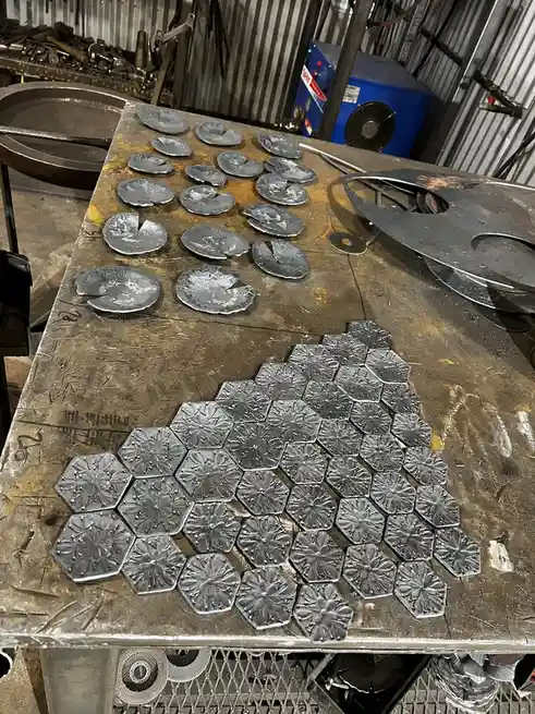 A cluttered metalwork bench displays two distinct sets of metal pieces: a pile of roughly shaped, irregularly cut circular…