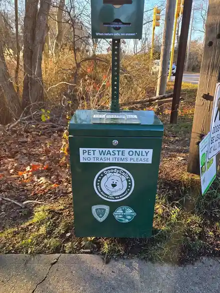 A green public pet waste disposal bin stands outdoors near a sidewalk, surrounded by fallen leaves and trees. It has a clear…