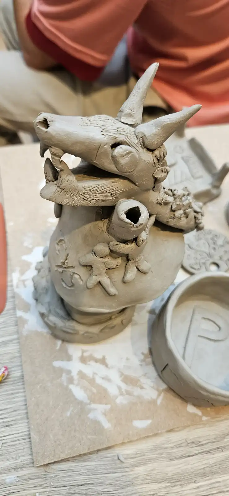 A close-up of a handmade clay sculpture depicts a dragon with two horns and an open mouth, appearing to bite into a small…
