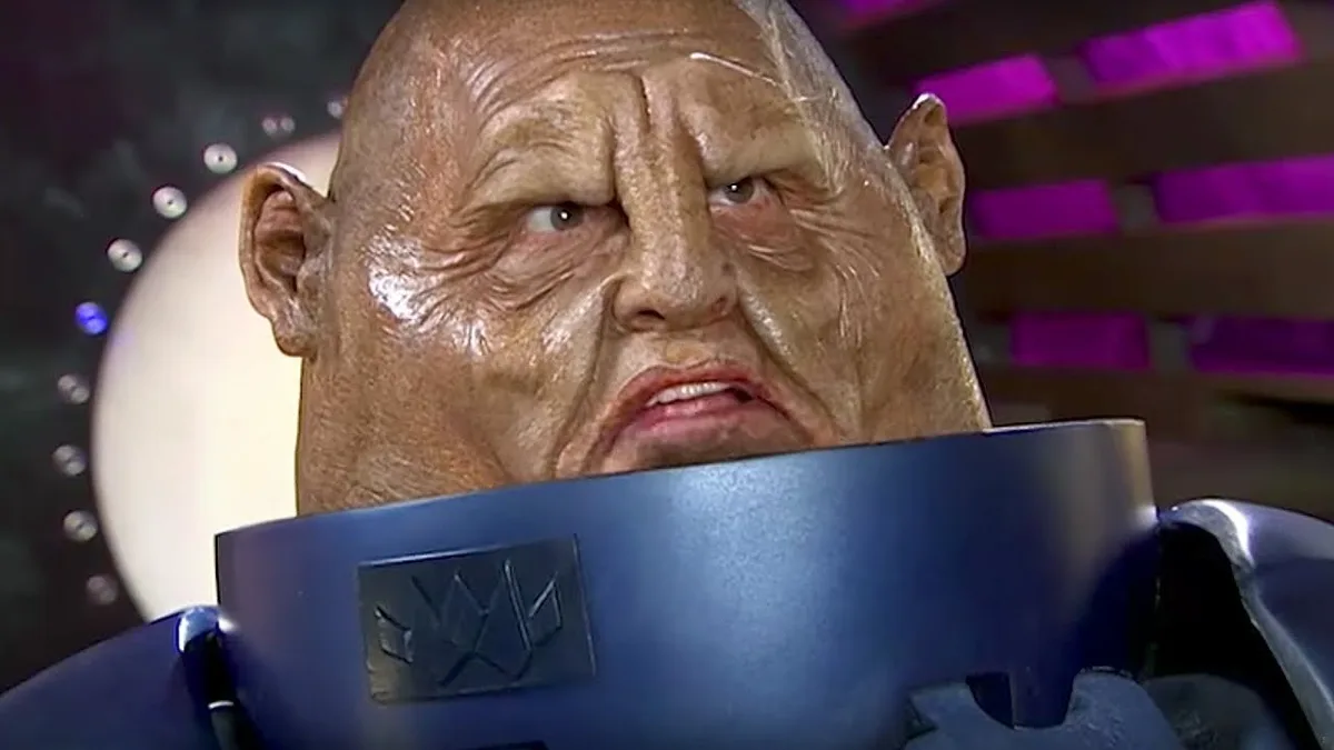 A close-up of a bald, wrinkled alien with a grimace, wearing a blue collar with a logo, against a purple-lit background. The…