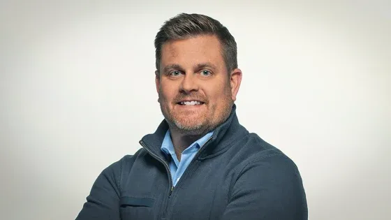 A man with short brown hair and a beard, wearing a dark blue zip-up sweater over a light blue collared shirt, smiles…