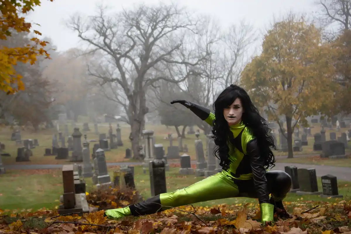 A person in a bright green and black superhero costume poses in a dramatic, low stance amidst fallen autumn leaves in a…