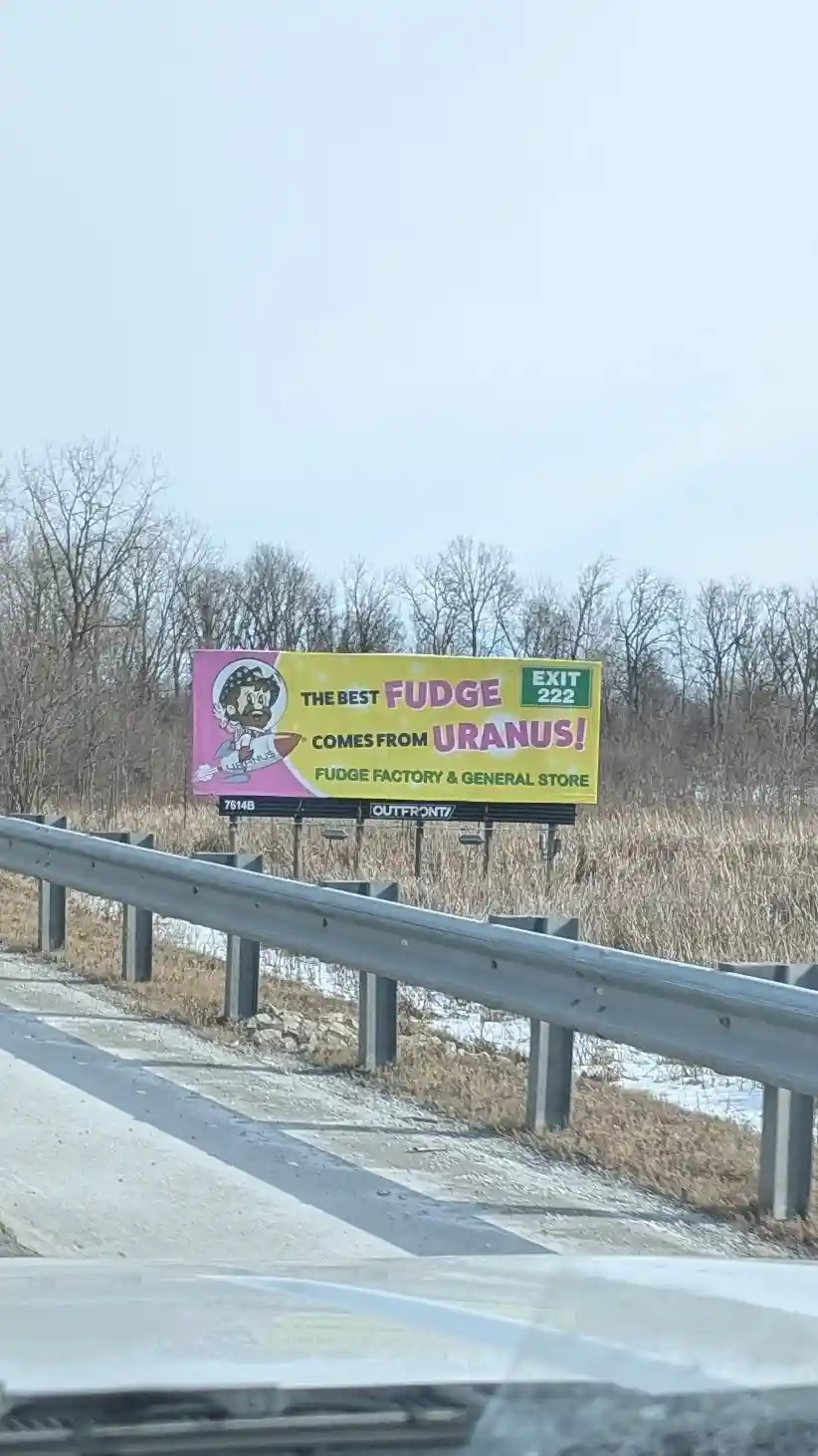 A roadside billboard viewed from a car, advertising 'THE BEST FUDGE COMES FROM URANUS!' with a cartoon character holding a…