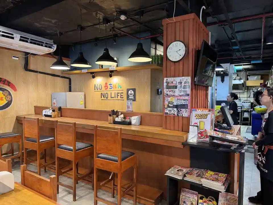 An interior shot of a Japanese-style restaurant with wooden bar seating and stools. On the back wall, colorful text reads…