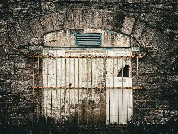 A weathered, rusty metal gate with vertical bars is set into a dark stone wall, partially obscuring a white door with a…