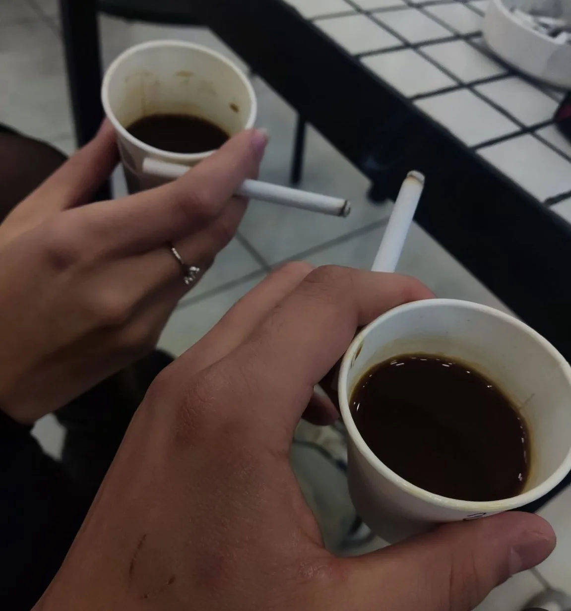 Two hands hold paper coffee cups with dark liquid, each with a cigarette balanced on the rim. One hand wears a ring. The…