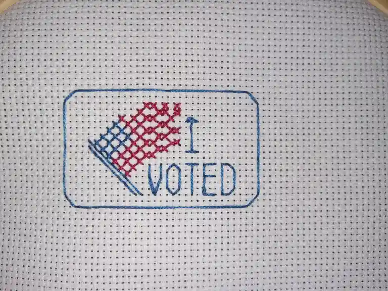 A close-up of a cross-stitch pattern on white fabric showing a rectangular design with blue outline, blue text reading 'I…
