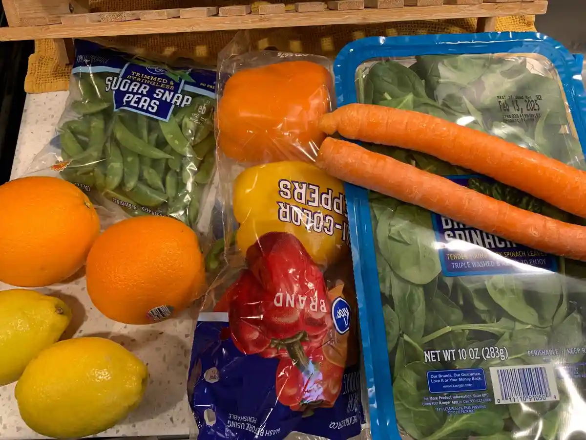 A grocery store produce display shows a variety of vegetables and fruits, including sugar snap peas, bell peppers, spinach…