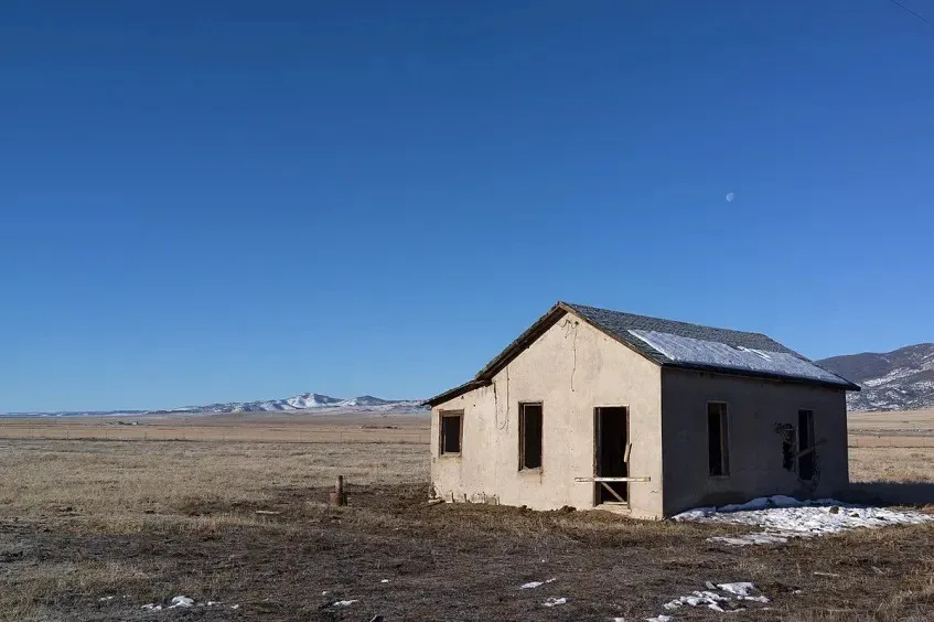 A dilapidated, windowless, doorless, and roof-damaged shack stands alone in a vast, barren field under a clear blue sky with…