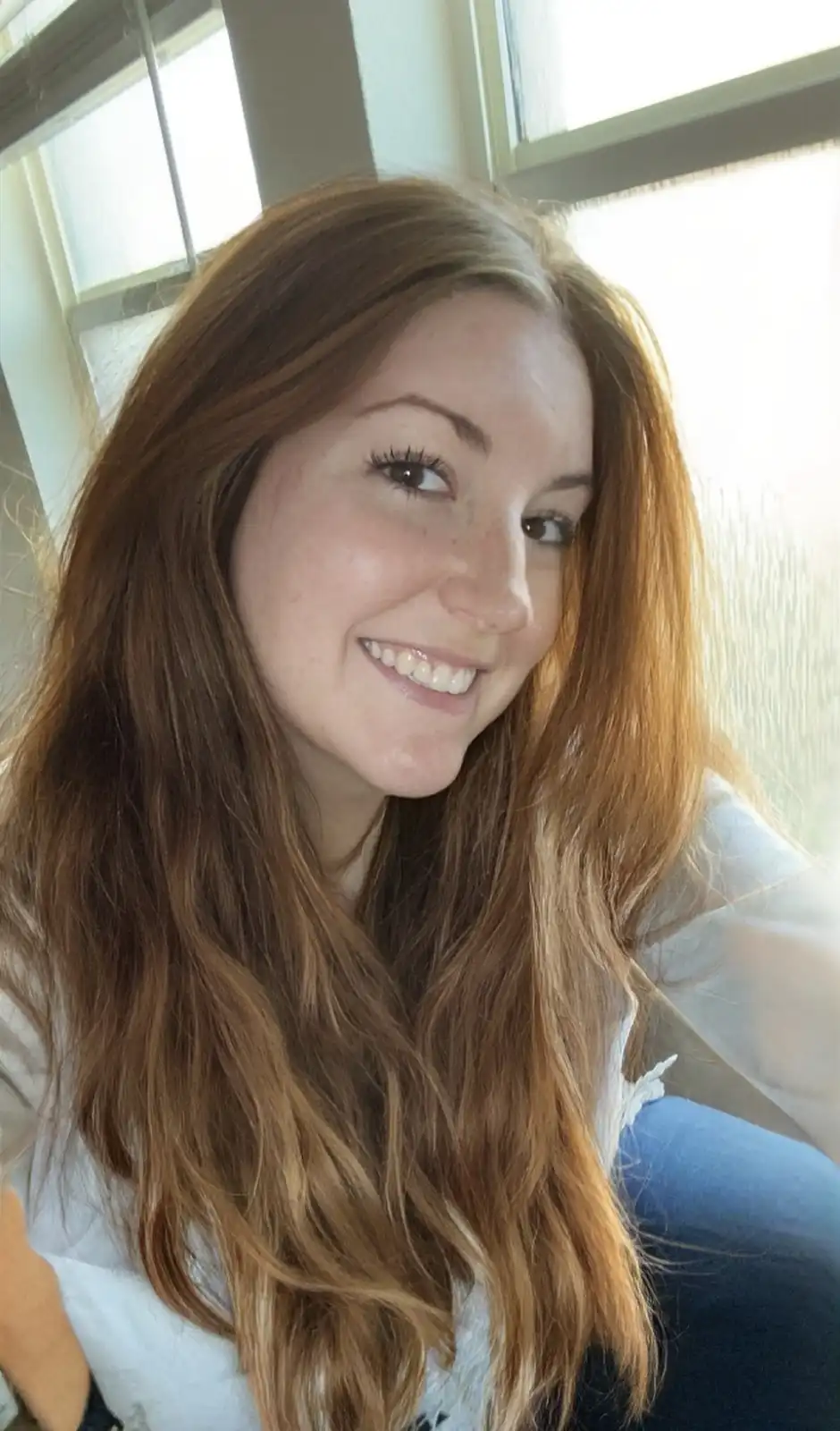A young woman with long, wavy reddish-brown hair smiles brightly at the camera while sitting near a window. She wears a…