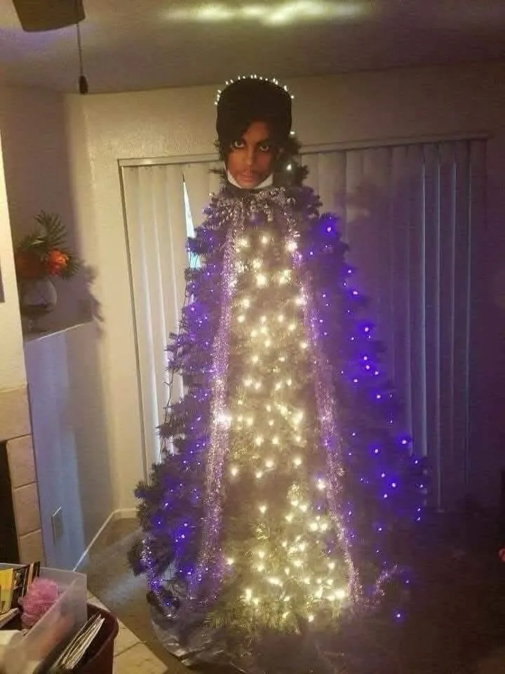 A person wearing a black wig and makeup resembling Prince stands inside a decorated Christmas tree, which is wrapped around…