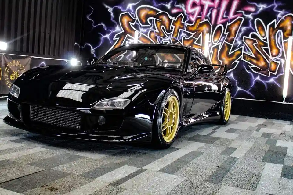 A glossy black Mazda RX-7 sports car with gold wheels is parked indoors on a checkered floor, facing the camera. Behind it…