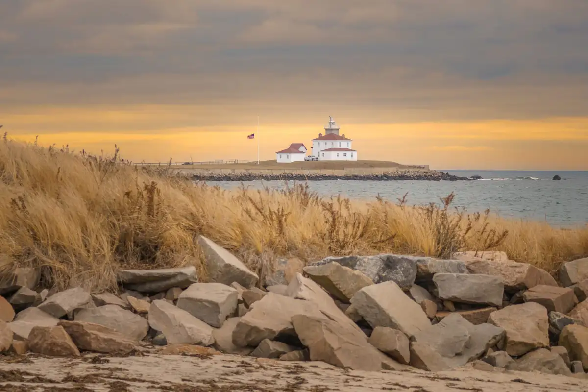 A scenic photo of a white lighthouse with red roofs on a rocky coastal headland at sunset, with golden grasses in the…