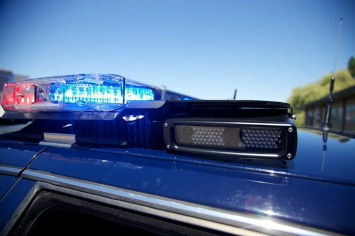 A close-up photo of a blue police car's roof with flashing red and blue emergency lights and a black camera mounted beside…