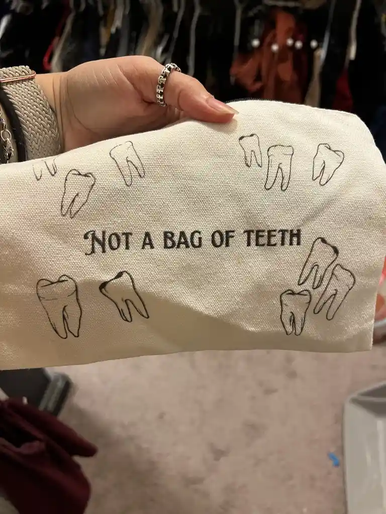 A hand holds a beige canvas pouch decorated with multiple black line drawings of teeth. The pouch has the text 'NOT A BAG OF…