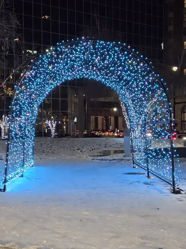 A nighttime photo of a large, arched structure covered in bright blue and white string lights, standing in a snowy urban…