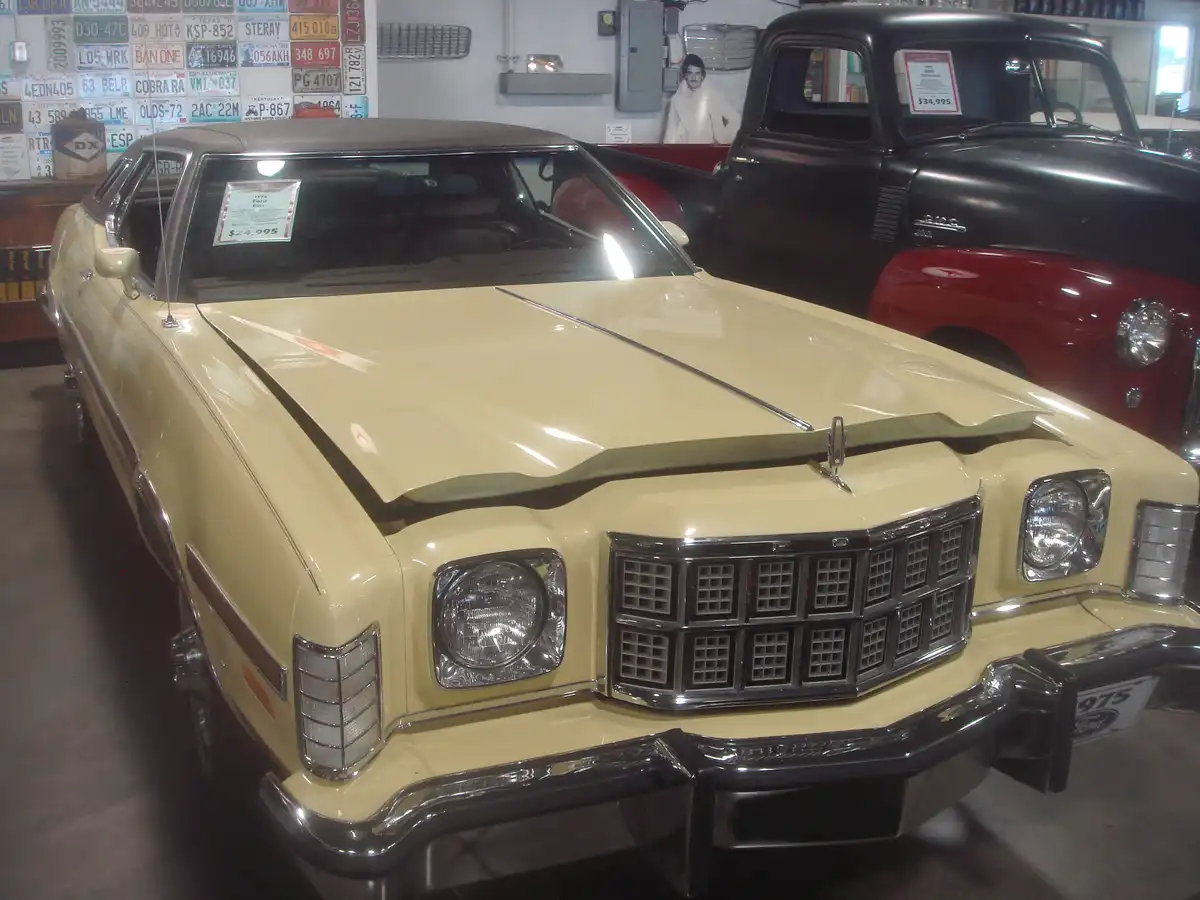 A beige 1975 Ford Elite is displayed in a car showroom with a price tag of $24,995 on its windshield. Behind it, a black and…