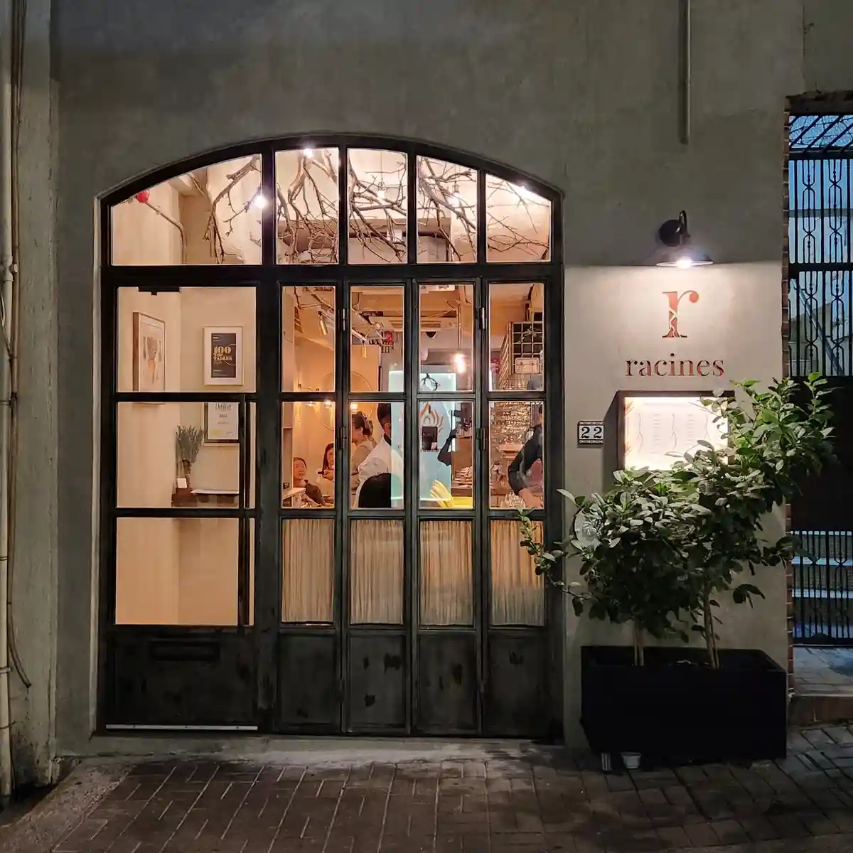 An exterior night shot of a restaurant named 'racines' with a large arched window and double doors, showing patrons dining…