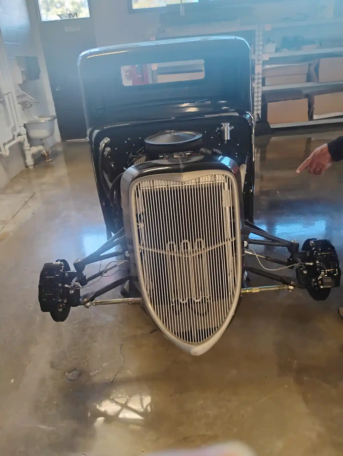 A vintage hot rod with its front end and engine exposed sits in a garage, missing its front wheels entirely. A person's hand…