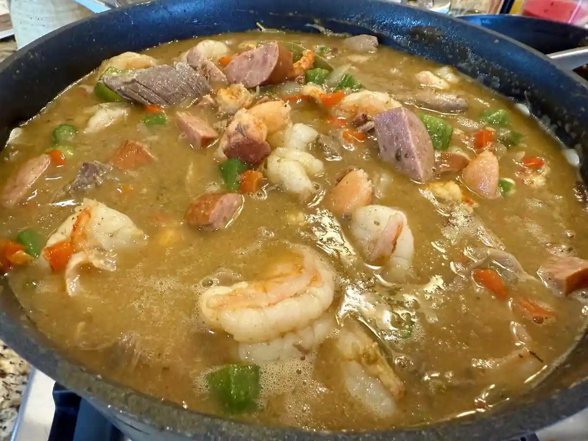 A close-up photo shows a bubbling pot of gumbo, a thick brown stew filled with shrimp, chunks of sausage, beef, green and…