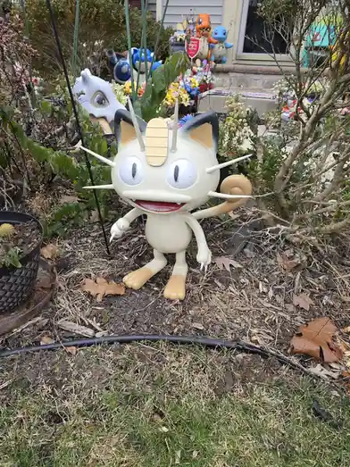 A large, cream-colored plastic statue of the Pokémon Meowth stands in a garden, its head and ears painted black, with a tan…