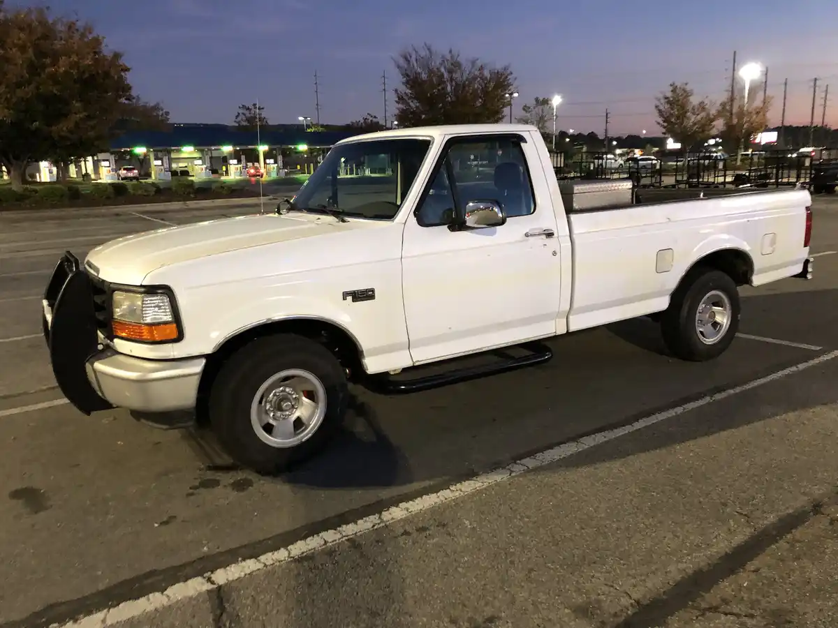 A white Ford F-150 pickup truck is parked in a parking lot at dusk, with a gas station visible in the background. The truck…
