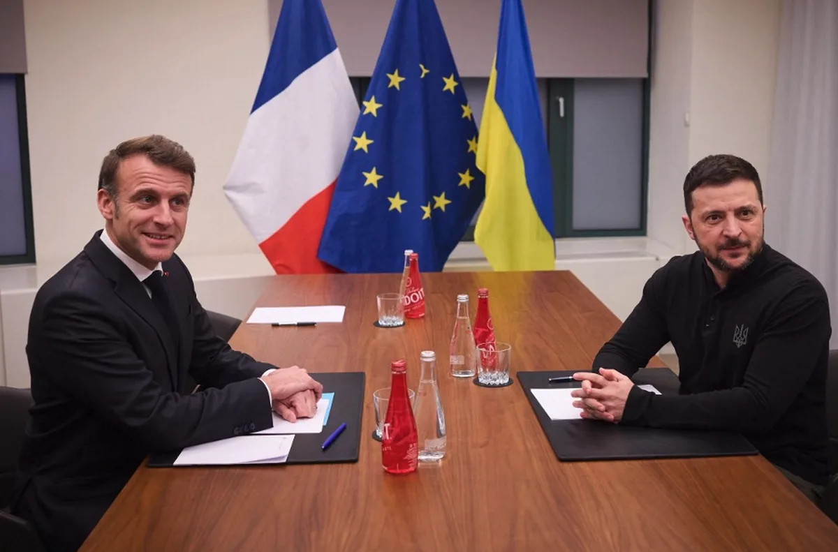 Two men, identified as French President Emmanuel Macron and Ukrainian President Volodymyr Zelenskyy, sit across a wooden…