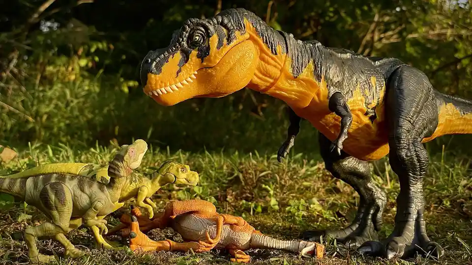 A large orange and black toy Tyrannosaurus Rex stands over a smaller orange toy dinosaur lying on the grass, while two…