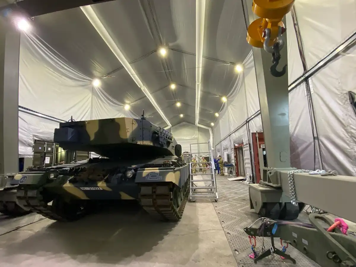 A large military tank with camouflage paint is parked inside a spacious white tent, surrounded by equipment and personnel…
