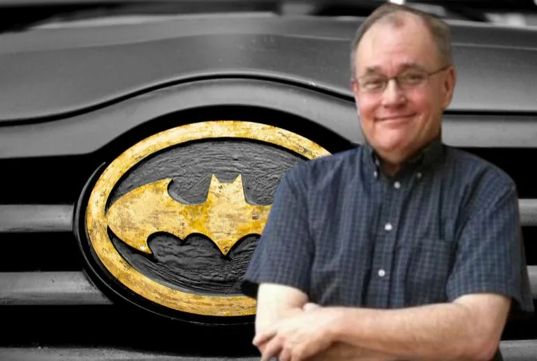 A man with glasses and a checkered shirt stands with arms crossed in front of a black vehicle. The vehicle's grille features…