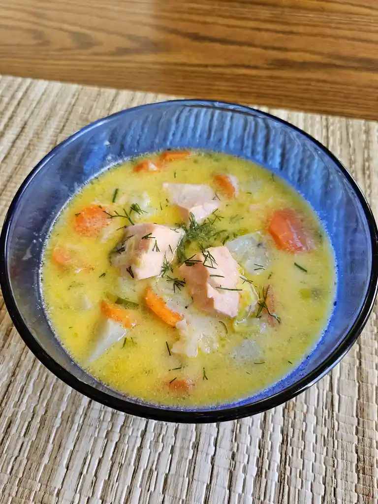 A top-down photo of a blue bowl filled with creamy yellow soup containing chunks of salmon, diced carrots, and chopped dill…