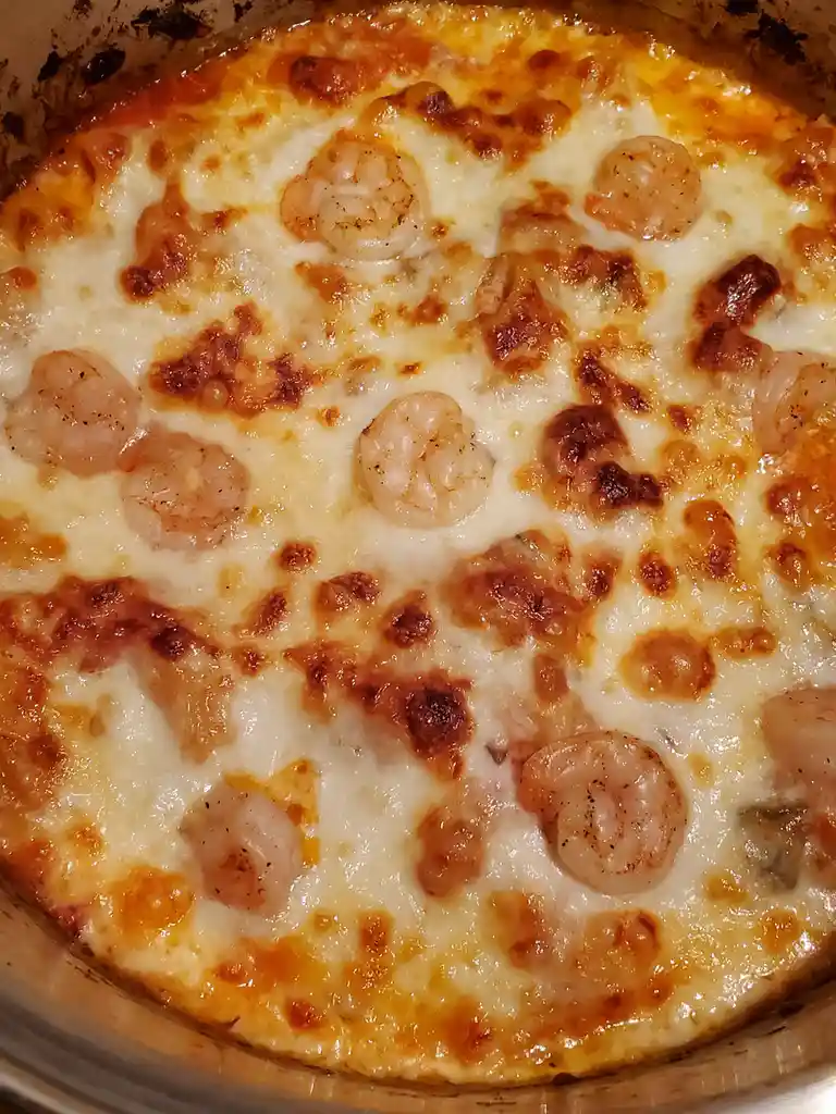 A close-up photo of a baked dish in a metal pan, featuring melted, golden-brown cheese with several cooked shrimp scattered…
