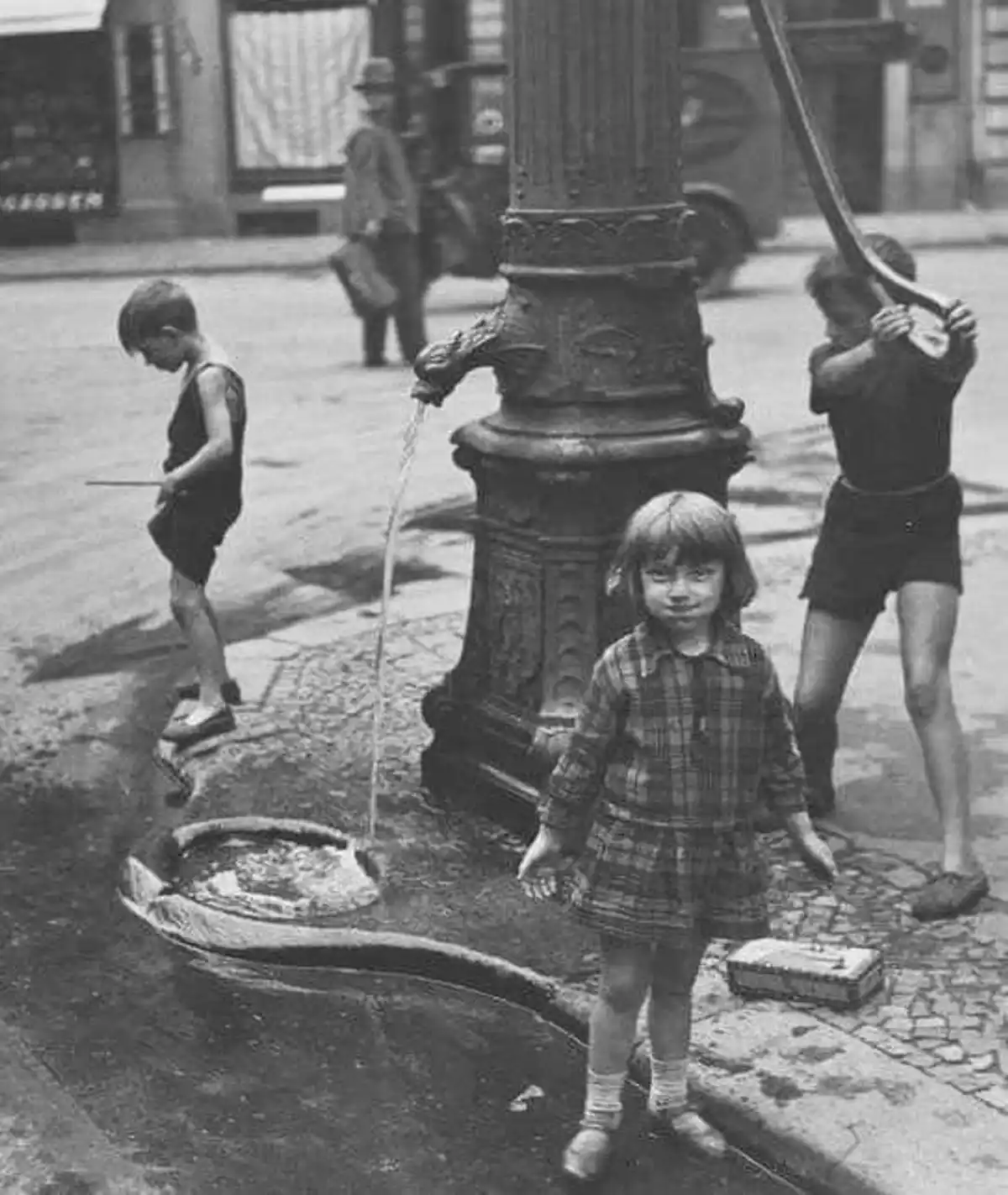 A vintage black-and-white photo shows three children around a public water pump on a cobblestone street. One child in a…
