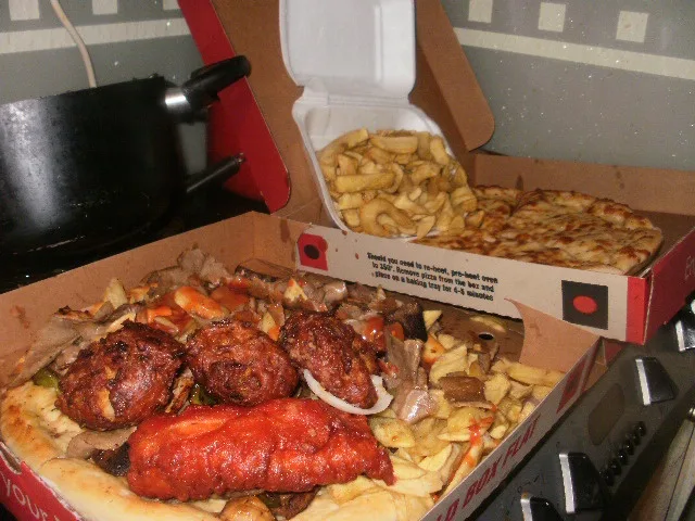 A messy, overflowing pizza box filled with fried chicken, ribs, and french fries sits on a kitchen counter next to another…