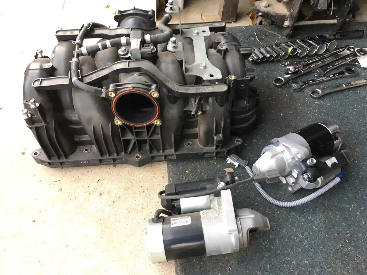 A disassembled black plastic automotive intake manifold sits on a garage floor next to two starter motors and a set of…
