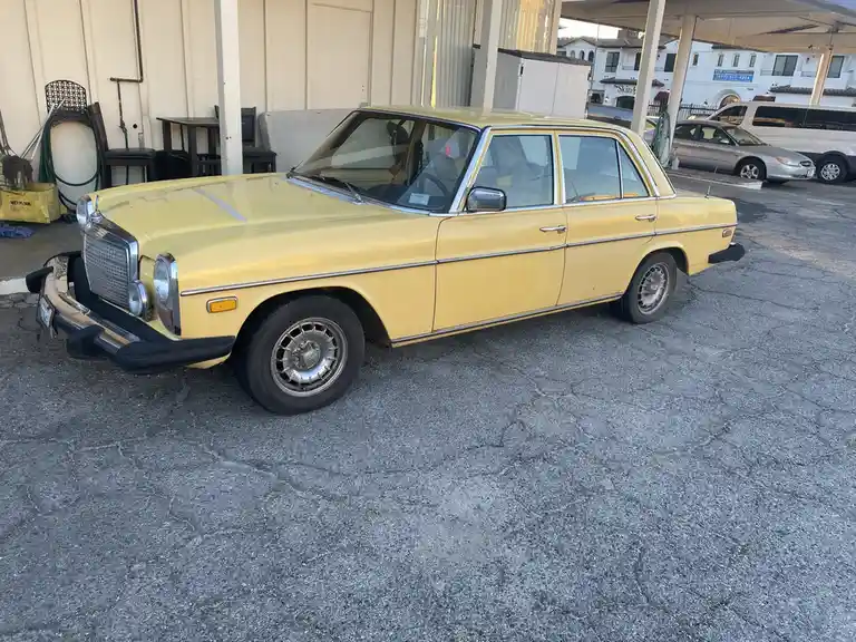 A pale yellow vintage Mercedes-Benz sedan is parked on cracked asphalt in front of a building. The car has chrome trim…