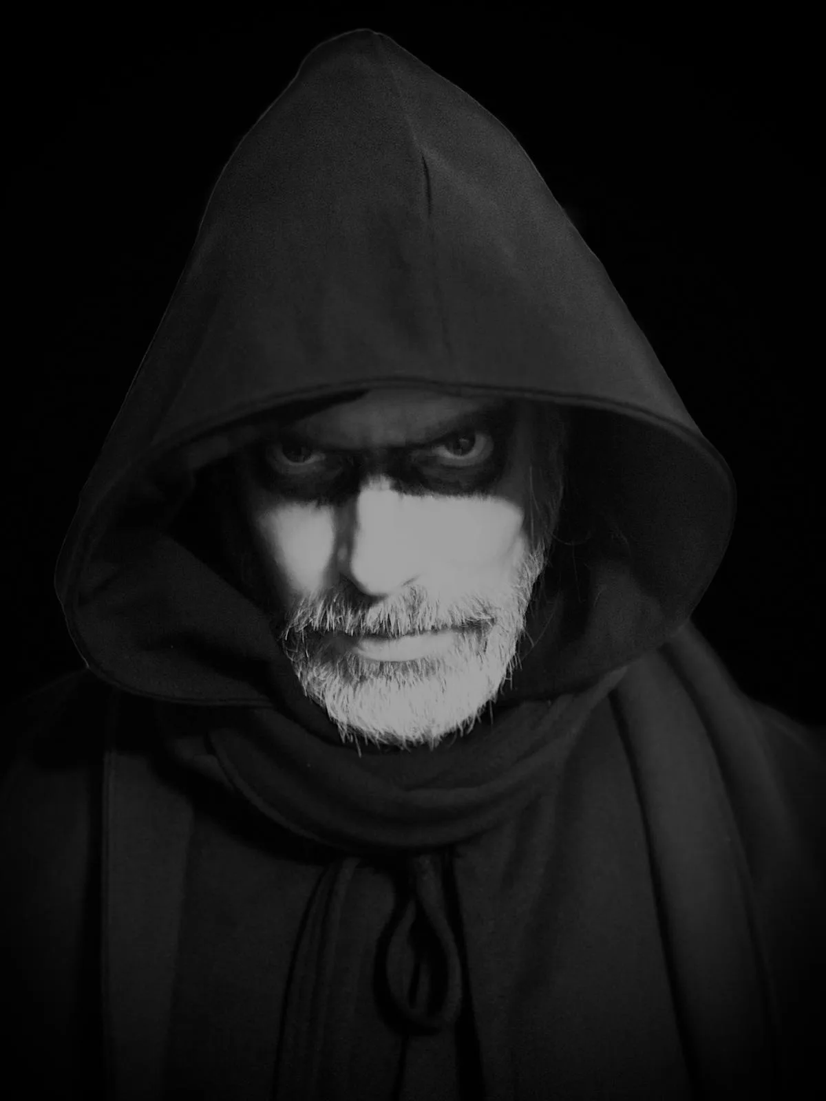 A black-and-white portrait of a man in a dark hooded robe, with stark white face paint around his eyes and beard, and dark…