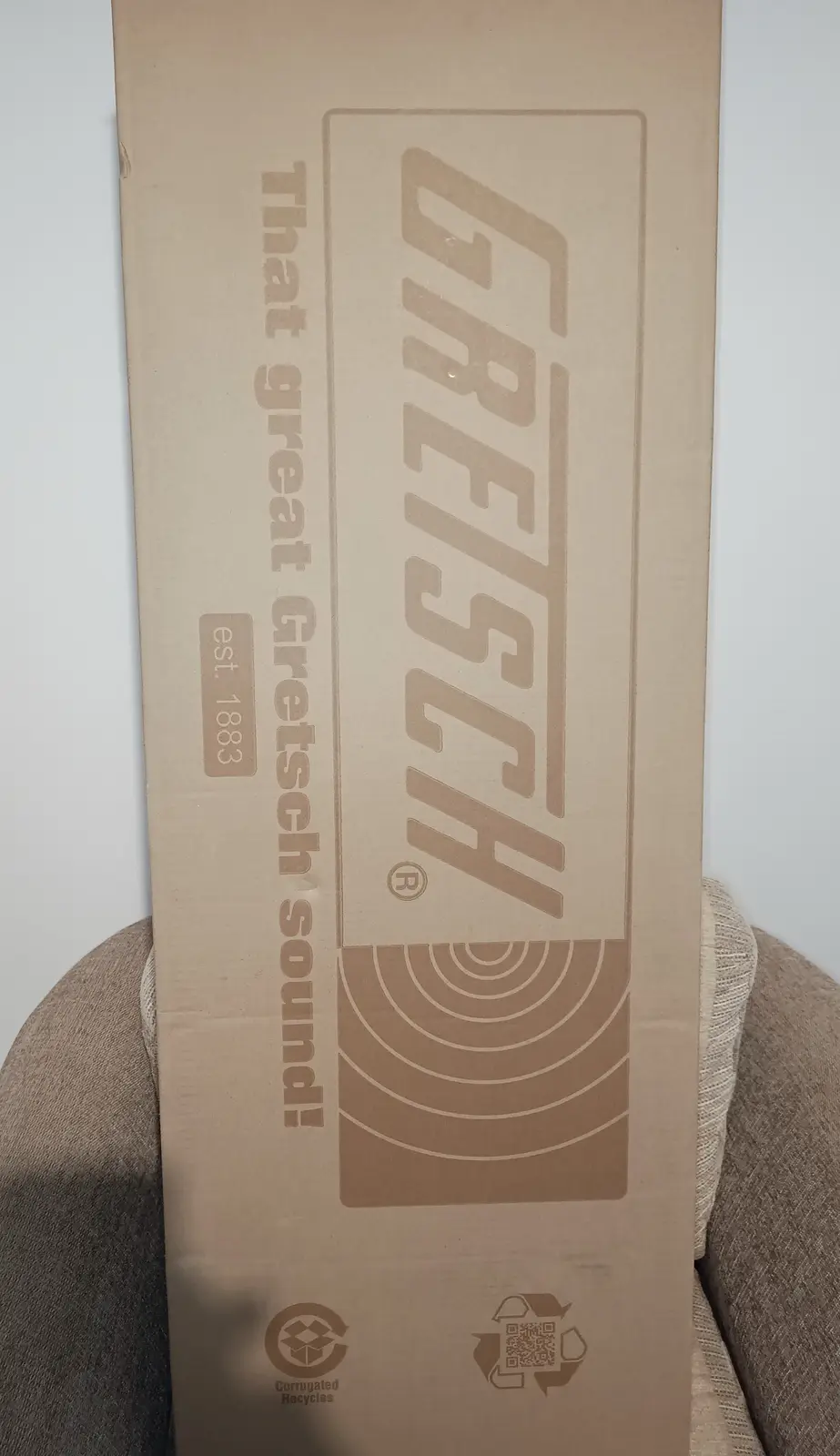 A tall, plain brown cardboard box, likely for a Gretsch guitar, stands upright on a textured gray fabric surface. The box…