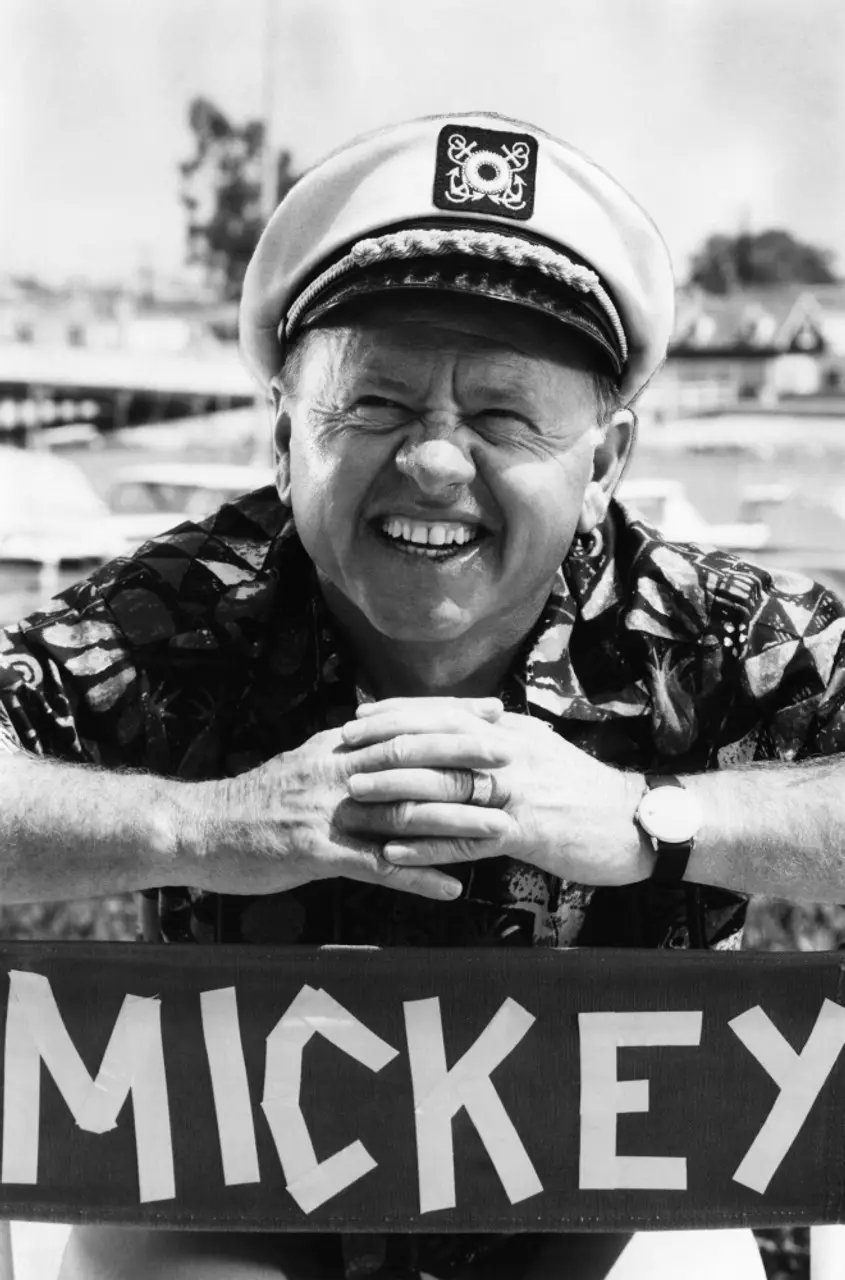 A black-and-white photo shows a smiling older man wearing a captain's hat and a patterned shirt, leaning on a railing with…
