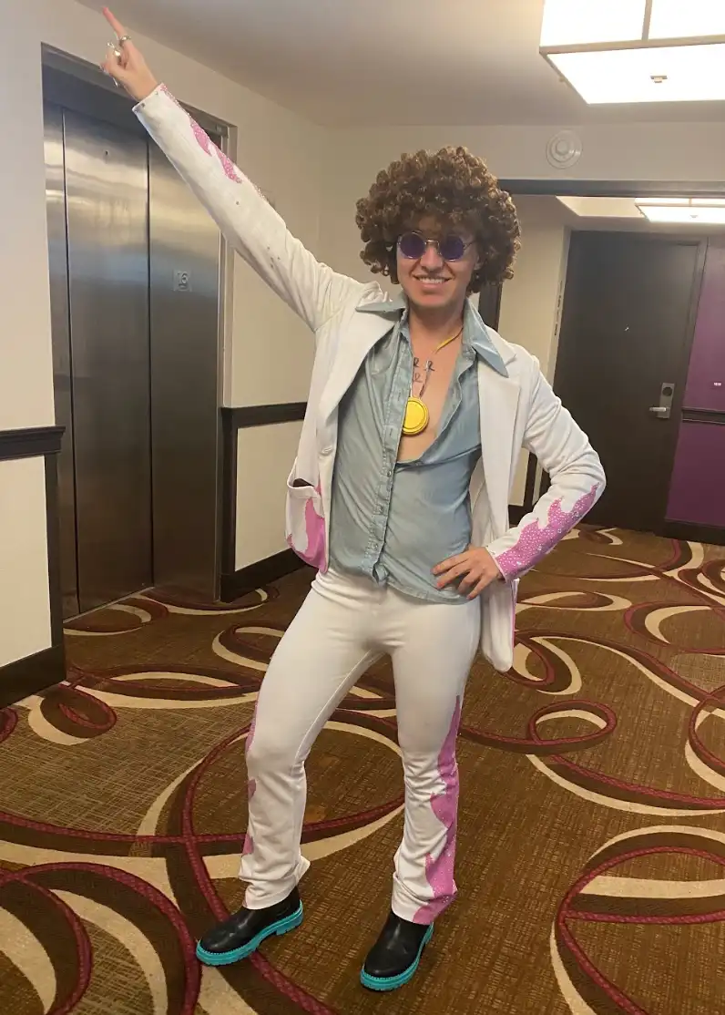 A person in a flamboyant 1970s-style outfit — including a curly wig, sunglasses, a white jacket with pink paint splatters…