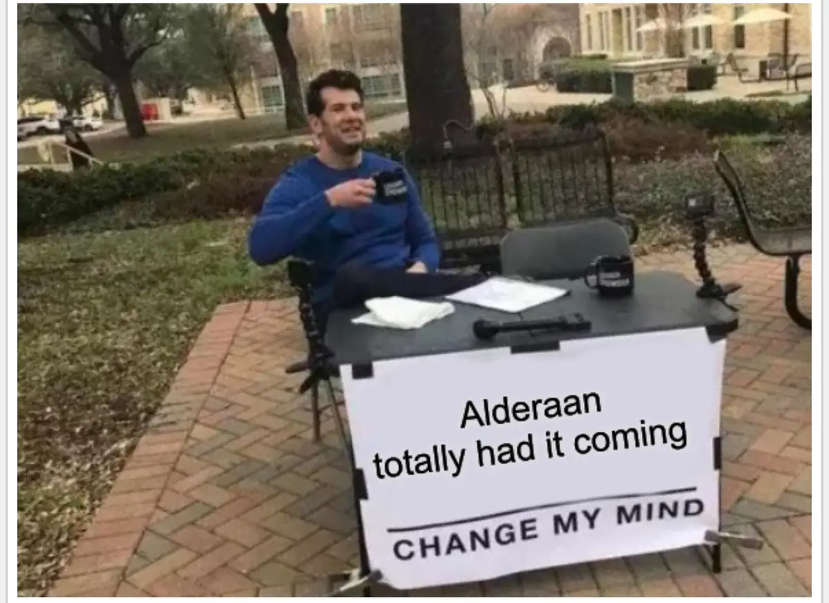 A man sits at a makeshift outdoor desk in a park, holding a mug, with a sign in front of him that reads 'Alderaan totally…