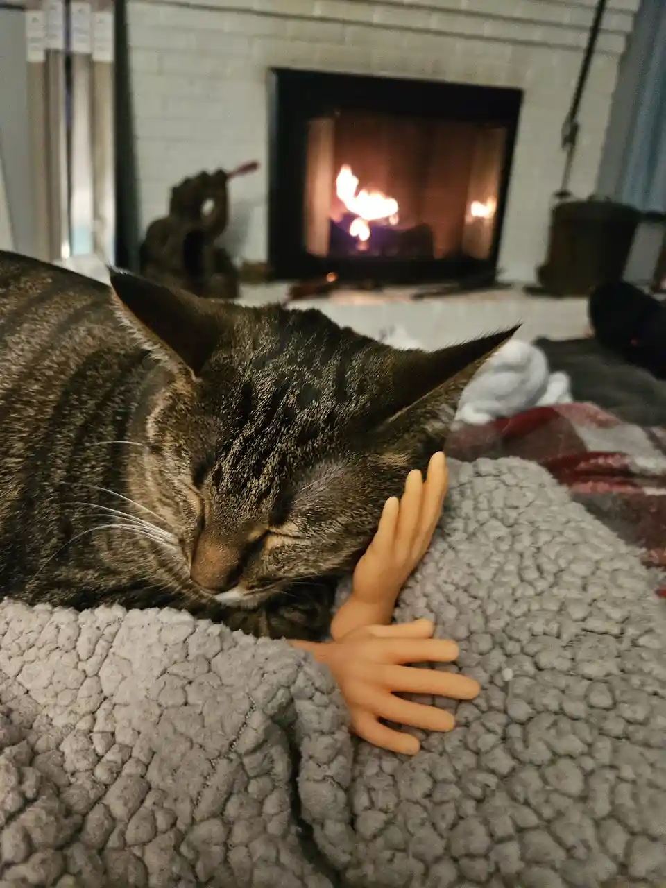 A tabby cat is sleeping soundly on a grey textured blanket in front of a lit fireplace, with two small plastic doll hands…
