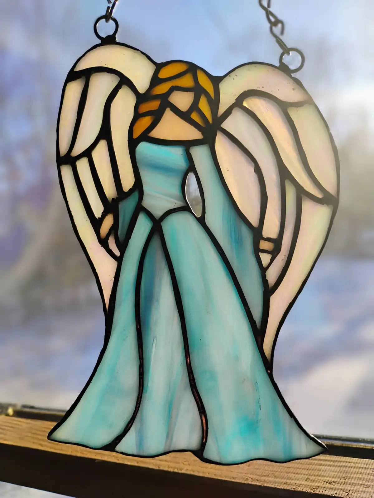 A stained glass angel ornament with large white wings and a blue robe hangs by chains against a snowy outdoor background…