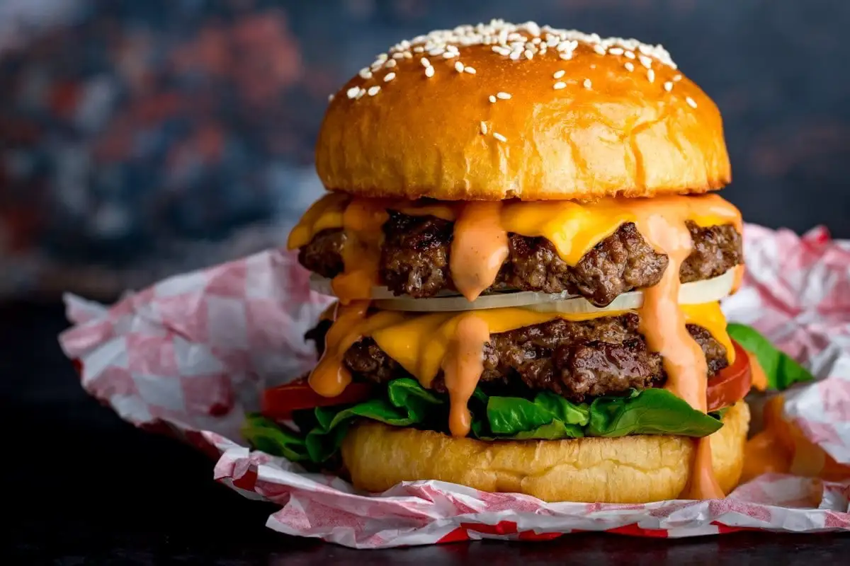 a visually appealing, tall double cheeseburger with two beef patties, melted yellow cheese, orange sauce dripping down…