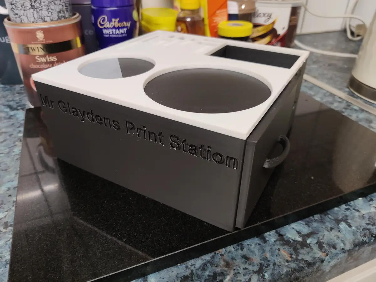 A 3D-printed black and white box labeled 'Mr Glaydens Print Station' sits on a kitchen counter, featuring two circular…