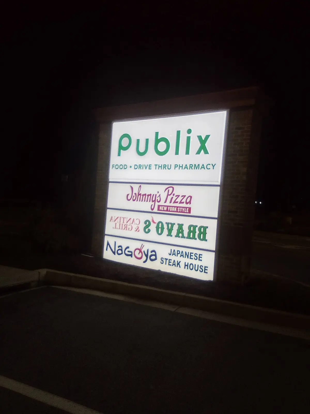 A nighttime photo of a brightly lit commercial sign for a Publix supermarket, which also advertises Johnny's Pizza, Bravo's…